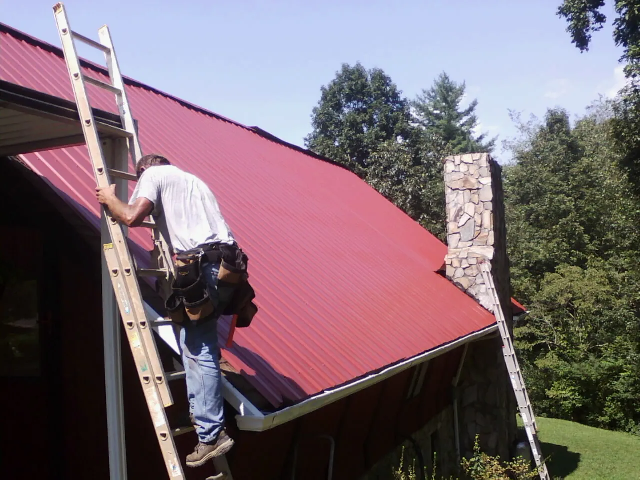 Roofer on a ladder beside a red metal roof and stone chimney — Soffit & Fascia Repair by Roofing Brothers in White Oak